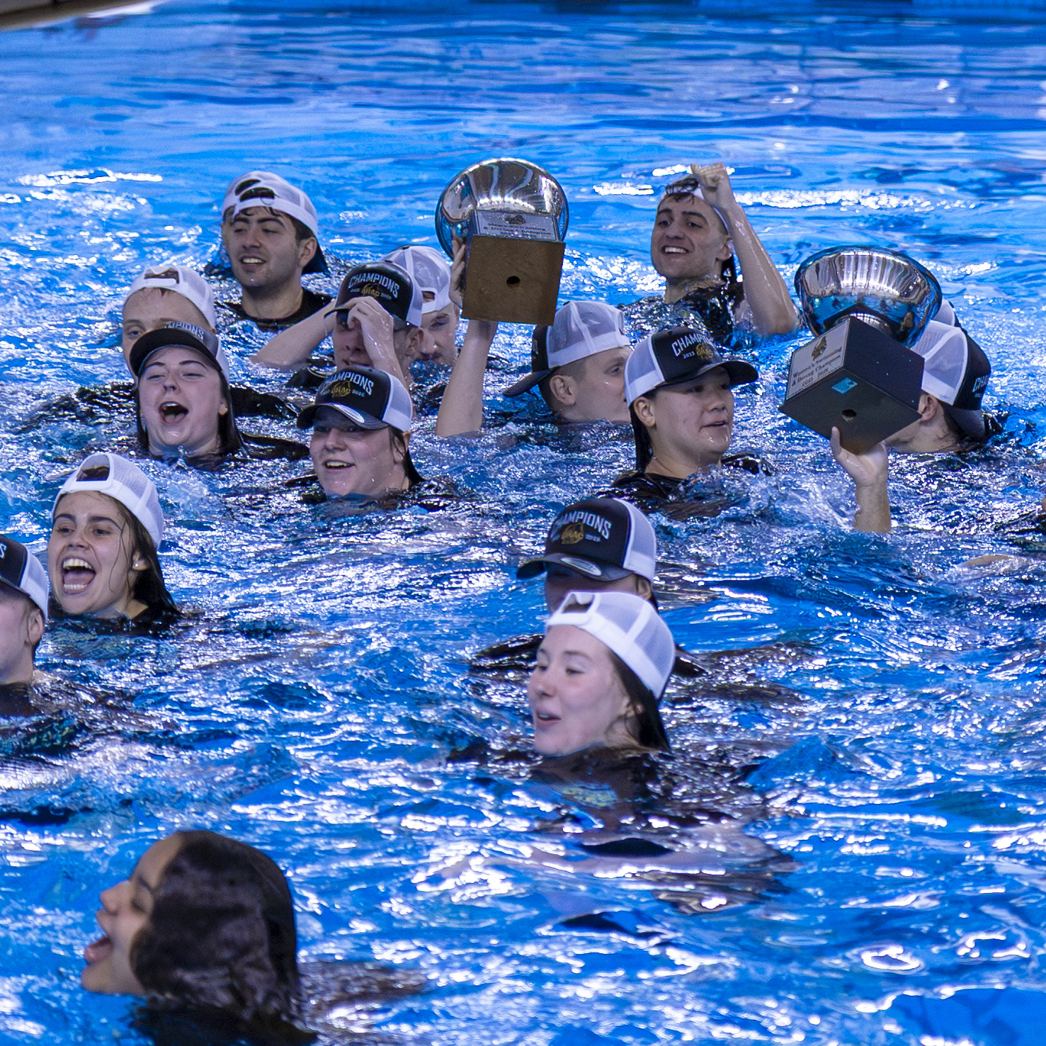 Swimmers in the pool wear matching hats and hold up trophies while gathered closely in the water.